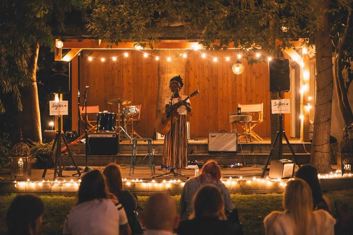 An outdoor wooden stage with a performer, string lights and an audience